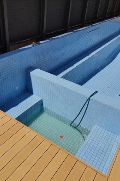 An empty rectangular swimming pool lined with small blue mosaic tiles, featuring a built-in spa area and surrounded by a light wooden deck