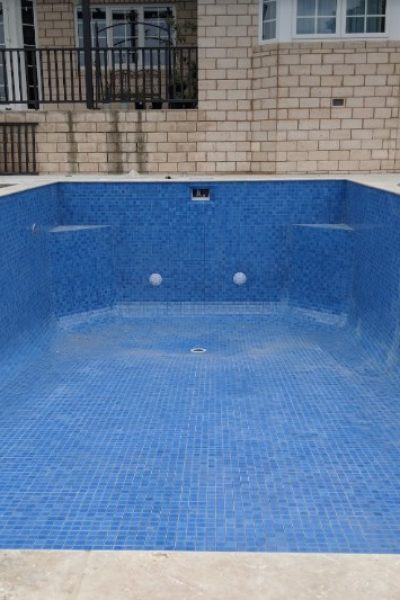 A free-form inground swimming pool with stone coping and a light mosaic tile waterline, partially filled with clear water